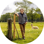 Carsten Bunk (ein weißer Mann mit mittellangen braunen Haaren und Brille) lehnt sich an den Zaun einer Alpaka-Weide
