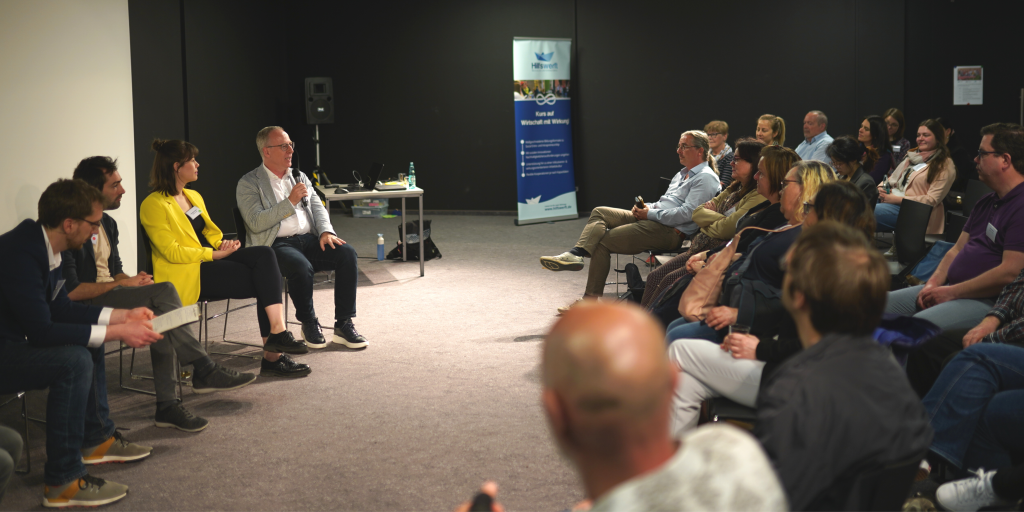 Dr. Christian Frühwald spricht auf unserer Podiumsdiskussion in ein Mikrofon. Er sitzt in einem Stuhlkreis neben Annegret Schnick, Marten Welschbach und Fabian Oestreicher. Gegenüber sitzt das Publikum des Praxis-Austausches in Stuhlreihen.