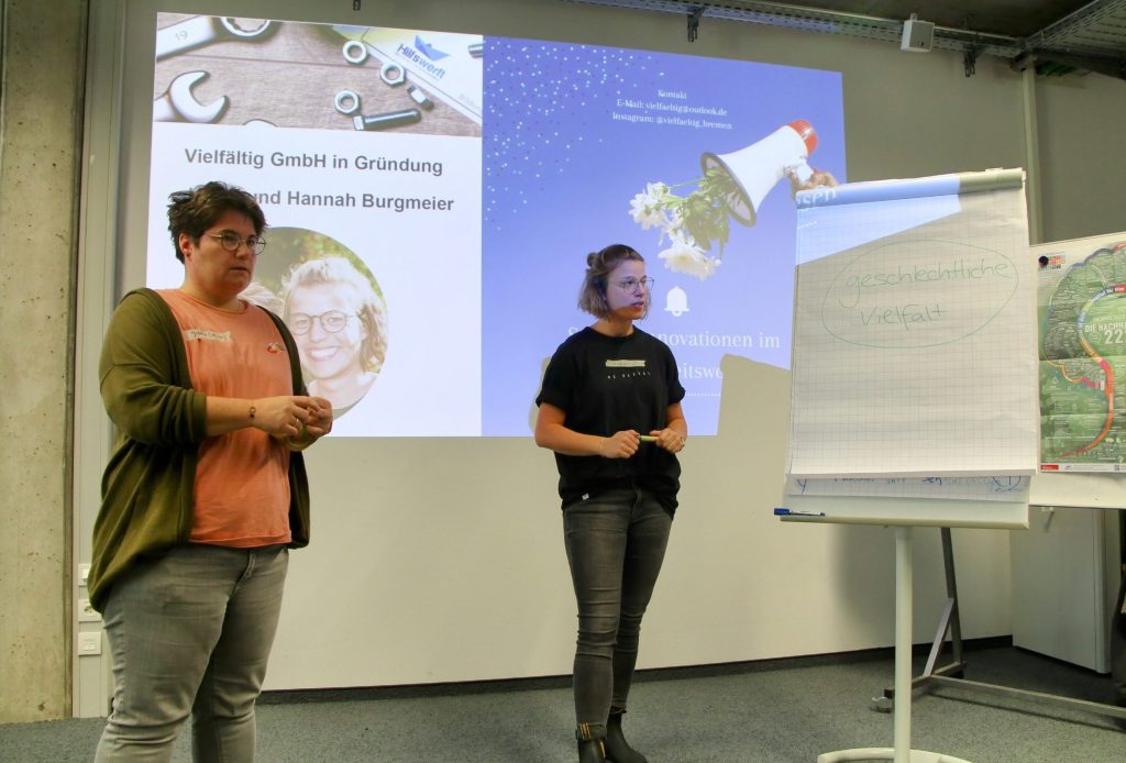Vor einer Leinwand mit dem Logo von vielfältig stehen Hannah und Judith Burgmeister und halten einen Vortrag. Auf einem Flipchart steht der Text "geschlechtliche Vielfalt".
