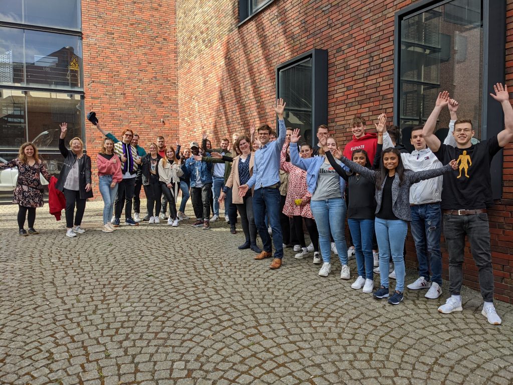 Vor einem Hochschulgebäude stehen ca. 25 junge Studierende und winken in die Kamera. Mit babei sind Referent Carsten Lessmann der Hilfswerft und Impulsgeberin Christine Sacher von den Elbe-Weser-Welten.