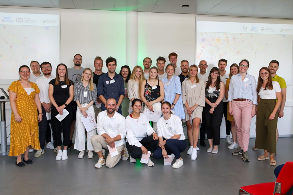 Vor einer beleuchteten Präsentationsleinwand stehen 24 Studierende der Uni Bayreuth gemeinsam mit Projektleiter Fabian Oestreicher und Professorin für Social Entrepreneurship Eva Jakob.